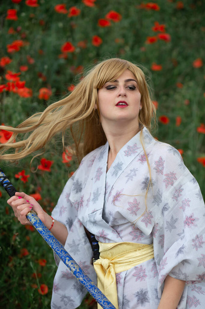 Young asian woman in traditional kimono trains fighting techniques with katana sword on the hills with red poppies, samurai warrior girl - Фото, изображение