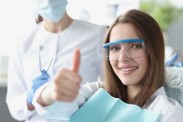 Молода жінка в захисних окулярах сидить у стоматологічному кріслі і показує великі пальці вгору
 - Фото, зображення