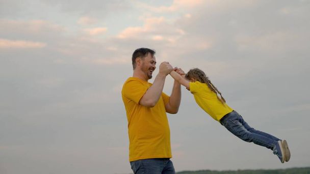 Isä leikkii, pyörittää onnellista tytärtään käsillään taivaan edessä kesäpuistossa. Isä ja pieni lapsi iloitsevat, nauravat ja halaavat yhdessä. Hyvää perhematkaa. Vauva on vanhempien käsissä. Onnellinen perhe - Valokuva, kuva