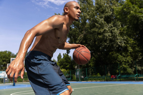 Close-up young man, african male basketball player playing basketball at street public stadium, sport court or palyground outdoors. Summer sport games. - 写真・画像