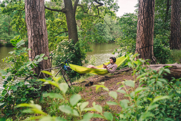 Joven hombre caucásico descansando en hamaca, escuchando música en los auriculares y usando el teléfono inteligente después en bicicleta en el bosque cerca del lago. Audio sanación. Lugar idílico. Viajar, acampar en la naturaleza. - Foto, Imagen