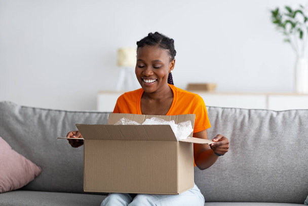 Online αγορές και παράδοση έννοια. Χαρούμενη μαύρη γυναίκα unboxing χαρτονένιο πακέτο, ικανοποιημένος με την αγορά στο σπίτι - Φωτογραφία, εικόνα