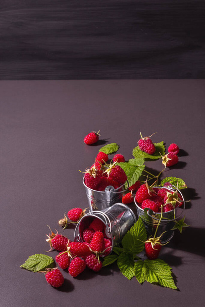 Ripe raspberries on a black stone concrete background. Small buckets, fresh leaves, new harvest. Trendy hard light, dark shadows, place for text - Fotografie, Obrázek