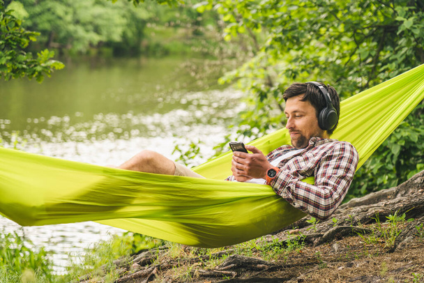 Buiten en avonturenconcept. Fietstocht naar het bos. Fietser rust in groene hangmat tussen bomen in de natuur door het meer tijdens het luisteren naar muziek op de hoofdtelefoon en surfen op internet op de smartphone. - Foto, afbeelding