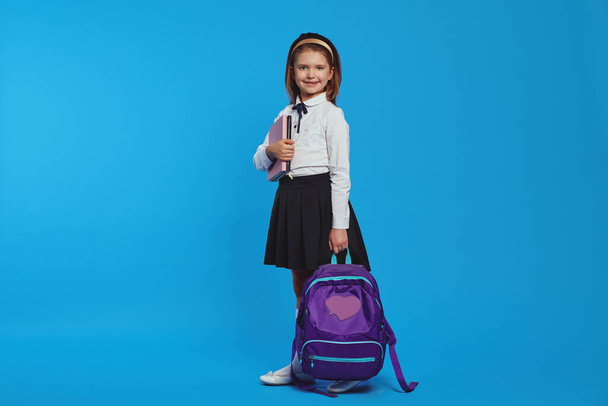 Horizontal shot of girl wearing backpack and school uniform, holding books - Photo, Image