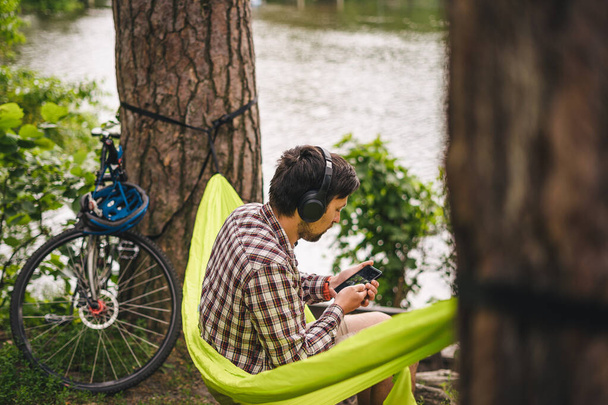 Young caucasian man resting in hammock, listening to music on headphones and using smartphone afterwards on bicycle in forest near the lake. Audio healing. Idyllic place. Travel, camping in nature. - Photo, Image