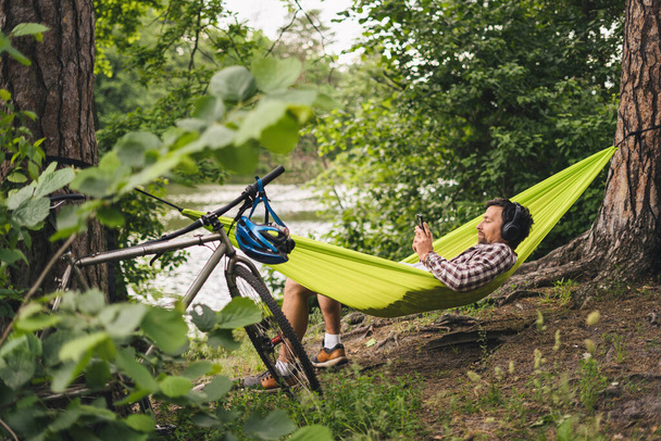Młody mężczyzna rasy kaukaskiej odpoczywający w hamaku, słuchający muzyki na słuchawkach, a następnie korzystający ze smartfona na rowerze w lesie nad jeziorem. Leczenie dźwięku. Idylliczne miejsce. Podróże, kemping w przyrodzie. - Zdjęcie, obraz