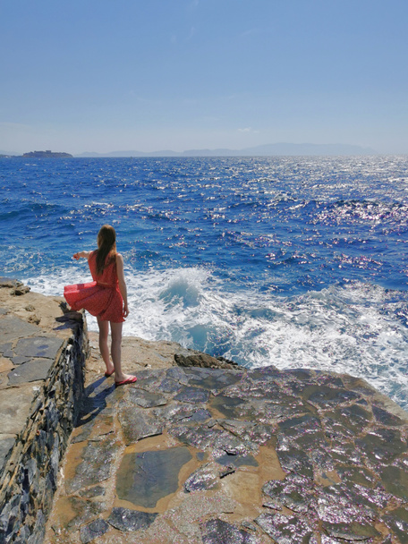 Egy gyönyörű vörös hajú lány vörös ruhában a hullámokat nézi a kék Égei-tenger hátterében. Törökország, Kusadasi. - Fotó, kép