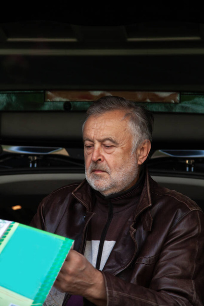elderly man with a gray beard in a leather jacket folds a road map against the background of an open trunk of a ca - Foto, afbeelding