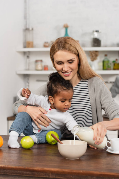happy woman pouring milk in bowl and holding adopted african american daughter  - Φωτογραφία, εικόνα