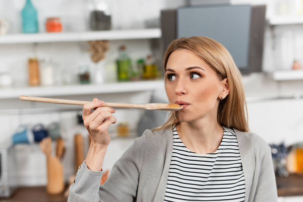 женщина держит деревянную ложку и пробует еду во время приготовления пищи на кухне  - Фото, изображение
