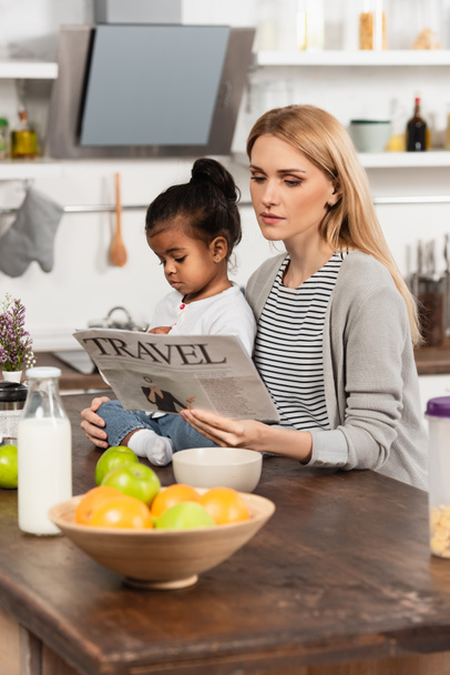 nő olvas utazási újság közelében örökbefogadott afro-amerikai lánya a konyhában - Fotó, kép