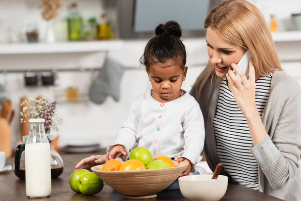 boldog anya beszél okostelefon közelében örökbefogadott afro-amerikai gyerek a konyhában  - Fotó, kép