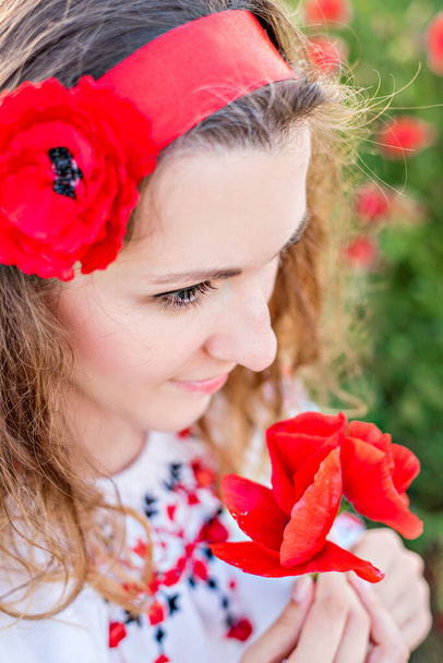 Close-up portrait of young woman in field, holding red poppy in hands. Girl in national costume, Headband with flower on head, an embroidered shirt and a red skirt. Nature walks in the countryside - Foto, imagen