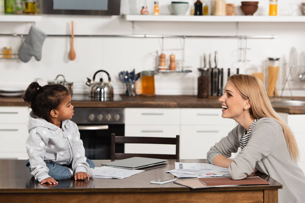 счастливая мать глядя на усыновленных африканских американская девушка на столе возле гаджетов  - Фото, изображение