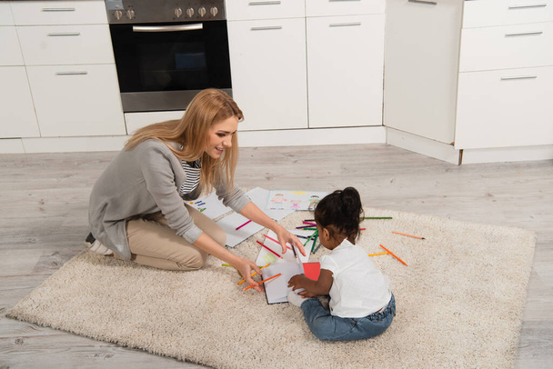 cheerful mother drawing with adopted african american girl while sitting on carpet  - 写真・画像
