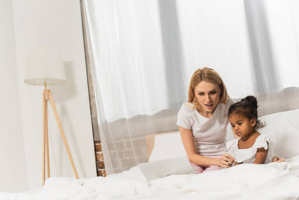 verbaasd moeder en geadopteerd Afrikaans Amerikaans kind kijken film op laptop in slaapkamer  - Foto, afbeelding
