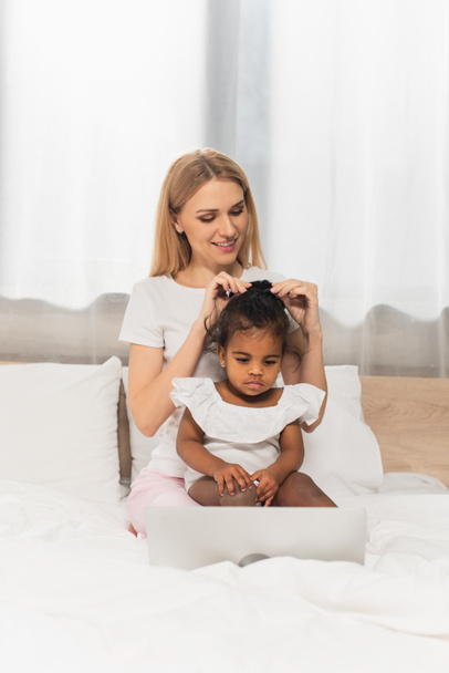 счастливая мать связывающая волосы усыновленного африканского ребенка, смотрящего фильм на ноутбуке в спальне  - Фото, изображение