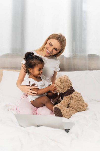 cheerful mother hugging adopted african american kid with teddy bear - Foto, imagen