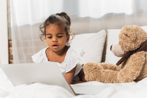 amazed african american kid watching movie on laptop near soft toy  - Фото, изображение