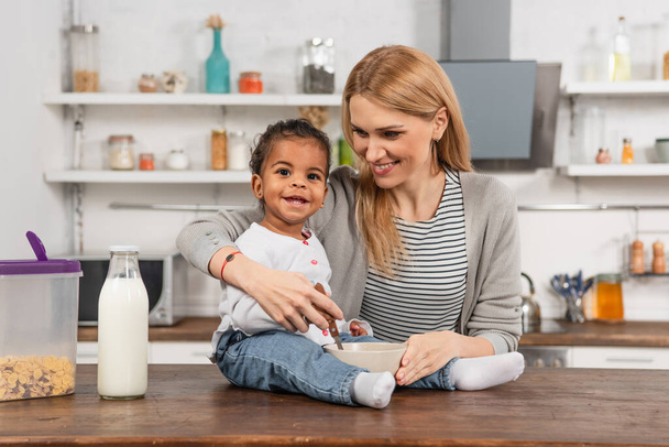 happy adopted african american kid holding spoon near bowl while sitting on kitchen table near smiling mother - Foto, afbeelding
