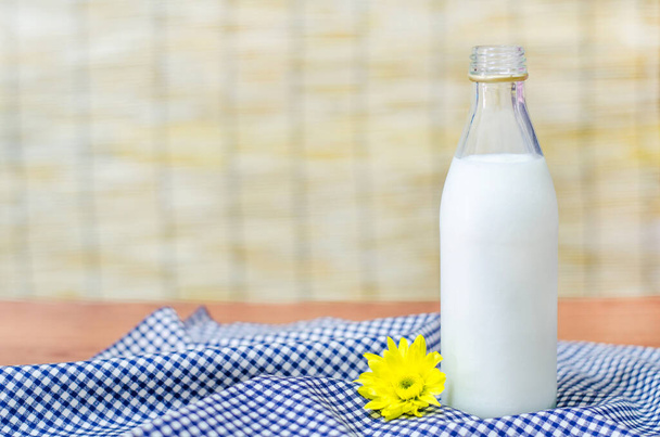Friss tej üvegben egy kis aranyos sárga virág fehér és kék asztali ruha felett elmosódott fa háttér - Fotó, kép