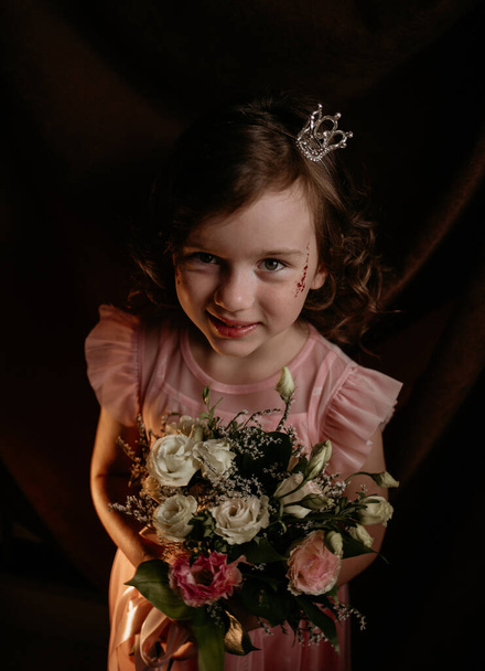 красива маленька кучерява принцеса з короною на голові тримає букет квітів на коричневому тканинному фоні
 - Фото, зображення
