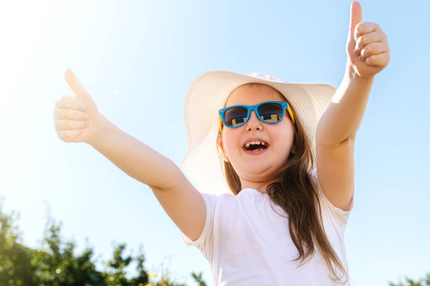 Güneş gözlüklü, beyaz tişörtlü ve mavi arka planda yaz şapkalı mutlu küçük kız. Yaz tatili, seyahat ve macera konsepti. - Fotoğraf, Görsel