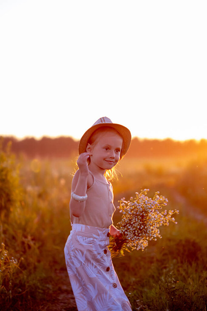 happy little caucasian girl 5 years old with a bouquet of flowers of chamomiles in the meadow looking at the camera, a portrait at sunset - Φωτογραφία, εικόνα