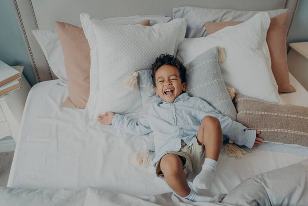 Afro-Amerikalı neşeli küçük çocuğun portresi. Yatakta yastıkların üzerinde gülüyor ve gülümsüyor. - Fotoğraf, Görsel