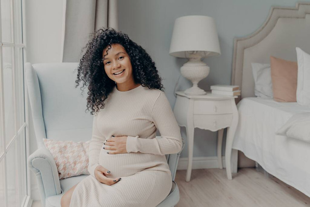 Young pregnant afro american woman in white dress posing in living room - 写真・画像