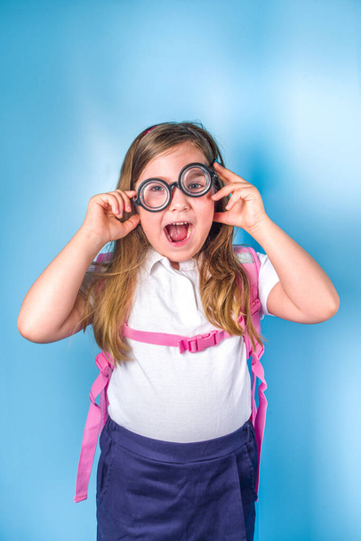 Вернемся к школьной концепции. Приглашение в школу, рекламный баннер. Милая ученица начальной школы в белой синей классической униформе, с книжным рюкзаком, смешными очками. Цветной синий фон - Фото, изображение