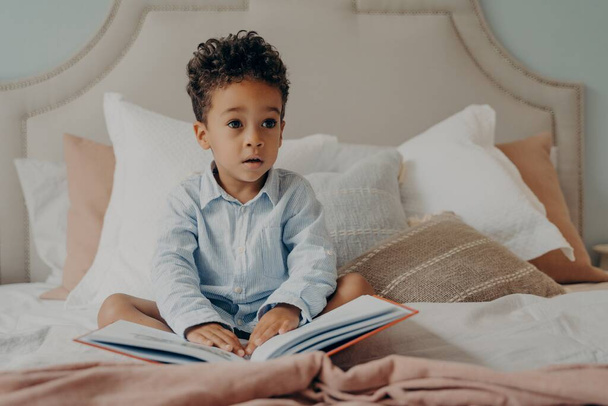 Cute curly haired afro american little boy sitting on bed with book - Foto, Imagem