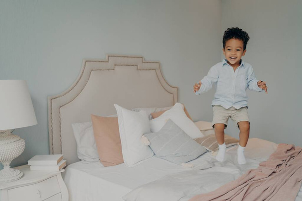 Happy beztroski mieszane rasy etniczne chłopiec w ubraniach casual zabawy i skakanie na łóżku - Zdjęcie, obraz