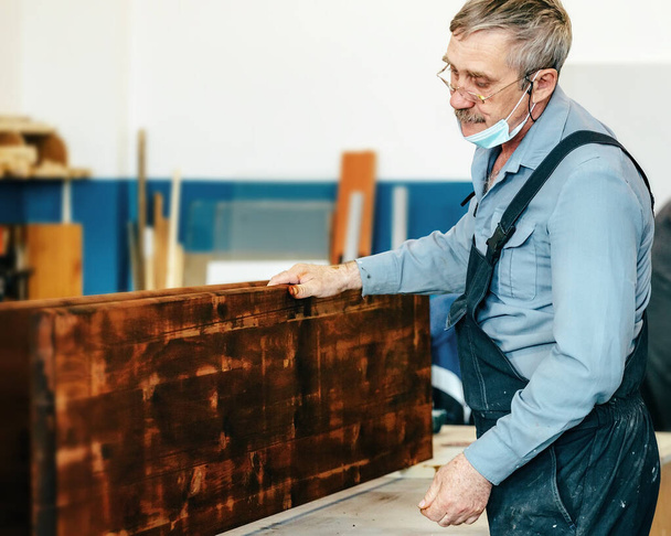 Um carpinteiro de cabelos grisalhos com óculos trabalha com madeira em uma bancada em uma loja de carpintaria.  - Foto, Imagem