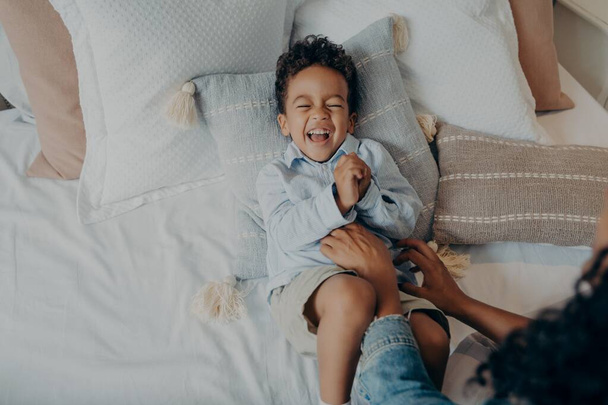 Suloinen pieni poika makaa tyynyillä sängyssä ja nauraa, kun äiti kutittaa häntä - Valokuva, kuva
