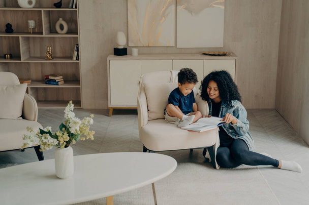 klein gemengd ras jongen lezen met zijn liefdevolle moeder terwijl genieten van tijd togerher in woonkamer thuis - Foto, afbeelding