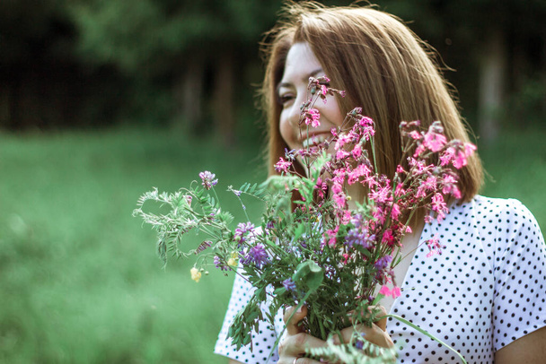A girl with a short haircut and flowers in her hands looks away and laughs. A close portrait of a young beautiful woman in a meadow with lilac wildflowers in her hands. Selective focus. Copy space. - Foto, immagini