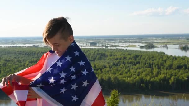 Chłopiec macha narodową flagą USA na zewnątrz nad błękitnym niebem na brzegu rzeki - Materiał filmowy, wideo