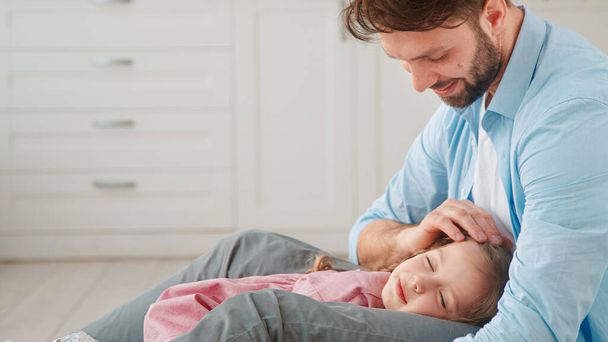 Papa souriant avec barbe regarde et caresse petite fille enfant avec de longs cheveux clairs dormir sur les genoux du père à la maison vue de près - Photo, image