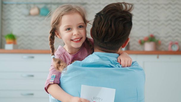 Милая маленькая девочка обнимает папочку в гостиной. Молодая семья в праздник Дня отца - Фото, изображение