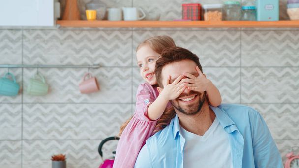 Dad and daughter spending time together at home. A little preschool girl covers her dad's eyes with her hands in the kitchen - Фото, изображение
