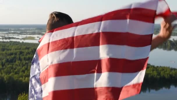 Chłopiec macha narodową flagą USA na zewnątrz nad błękitnym niebem na brzegu rzeki - Materiał filmowy, wideo