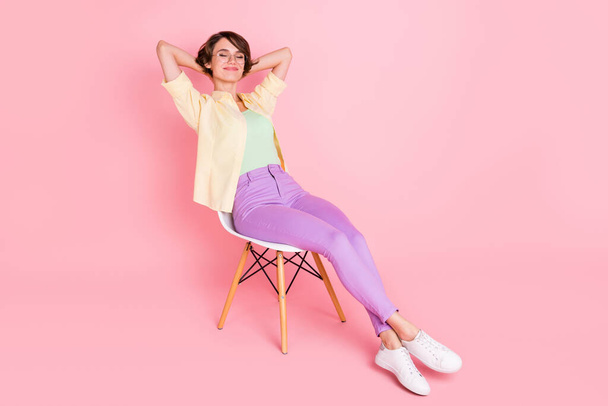 Full size photo of optimistic brunette lady sit on chair arms behind head wear shirt trousers sneakers isolated on pastel pink background - Φωτογραφία, εικόνα