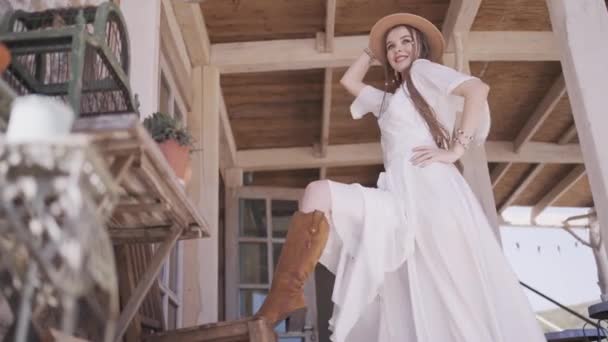 Rapariga do campo no alpendre, vestido branco e chapéu de cowboy. Acção. Visão inferior de um modelo feminino posando com a perna na bota de cowboy marrom em uma mesa de madeira. - Filmagem, Vídeo