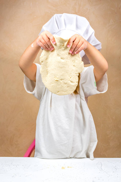Ein kleines Mädchen hält den Teig nach Gewicht, bedeckt ihr Gesicht. Die Entwicklung der Feinmotorik bei Kindern. Salzteig-Modellierung. - Foto, Bild