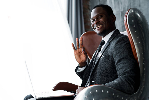 Photo from the side. Happy african american businessman working on laptop and showing okay sign smiling - 写真・画像