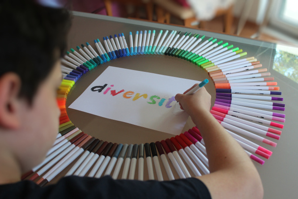 Młody chłopiec namalował słowo "różnorodność", którego prześcieradło znajduje się w środku dziesiątek długopisów pędzelkowych wszystkich kolorów. Reprezentuje równość, integrację, płeć, szacunek, tolerancję, orientację seksualną - Zdjęcie, obraz