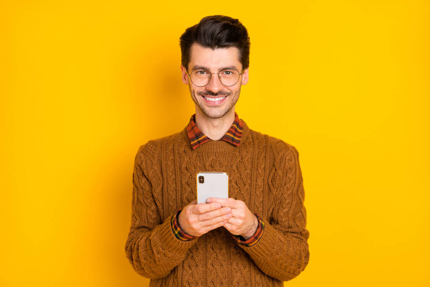 Photo of positive handsome guy hold telephone beaming shiny smile wear brown sweater isolated bright yellow background - 写真・画像
