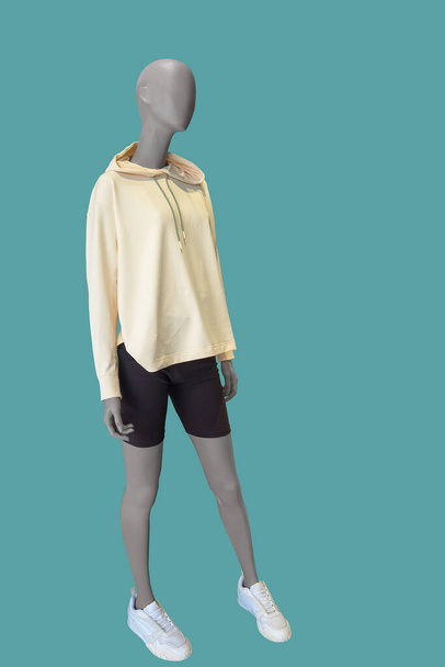 Imagem de comprimento total de um manequim de exibição feminino usando camisola com capuz amarelo e shorts pretos isolados em um fundo azul. - Foto, Imagem
